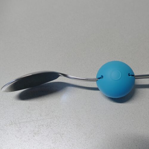 Um den Griff eines Eßlöffel ist ein blauer Ball gesteckt. Dieser hat einen Durchmesser von circa 5 cm und mittig einen Schlitz.