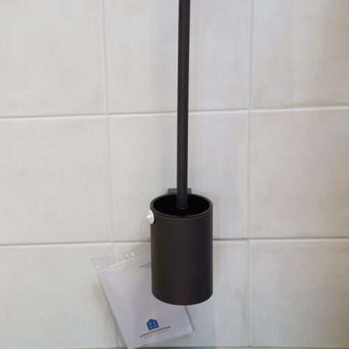 An der Wand befestigt. Schwarzer Halter in Säulenform, Durchmesser circa 7 cm, Höhe circa 15 cm, in dem eine schwarze WC-Bürste steckt.