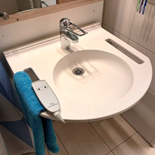 Das Waschbecken hat links und rechts Grifflöcher. Links ist ein blaues Handtuch eingehängt. An der Vorderseite ist die Form ein Kreissegment. Die kabelgebundene Fernsteuerung liegt oben auf.