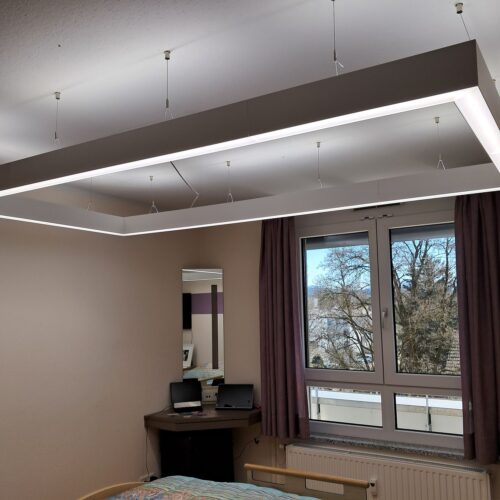 Der Rahmen der Leuchte ist fast so groß wie das Bett, das sich darunter befindet. Das Lichtband darauf ist circa 5 cm breit. Hier leuchtet ist in einem Weißton.