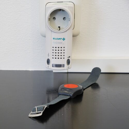 Der Empfänger mit dem Lautsprecher steckt in der Steckdose. Davor liegt der Sender in der Optik einer Uhr mit rotem Signalknopf.