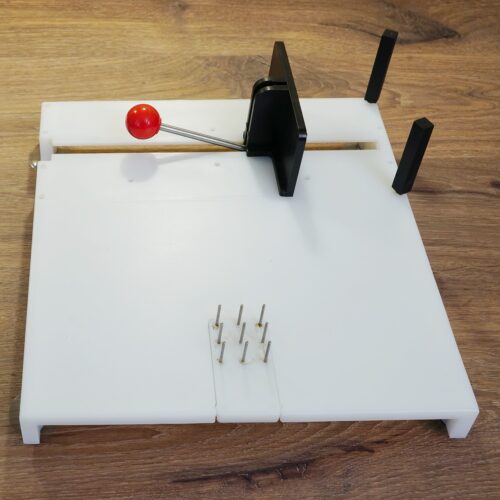 Weißes Kunststoffbrett mit einer Fixierfläche aus hochstehenden Nägeln (9 Stck ca. 1 cm hoch), und zwei circa 5 cm hohen Pfeilern, denen die der zu haltende Gegenstand mit enem Schieber geschoben werden kann.